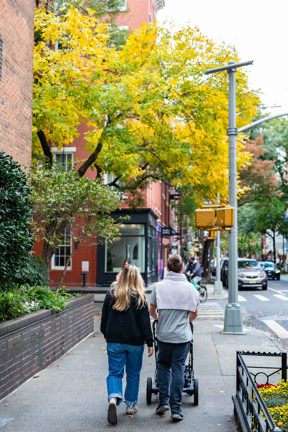 A couple walking on a sidewalk and pushing a baby stroller in the West Village as the tree above them begins to yellow for autumn, one of the best places to live in New York City.