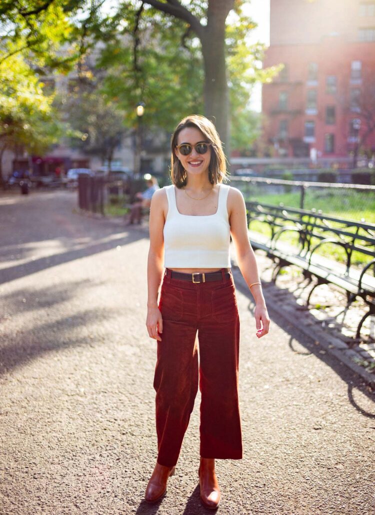 Smiling woman wearing a white tank top and maroon wide-legged pants, the perfect May outfit for NYC enjoying the sunshine at Tompkins Square Park with empty benches in the background