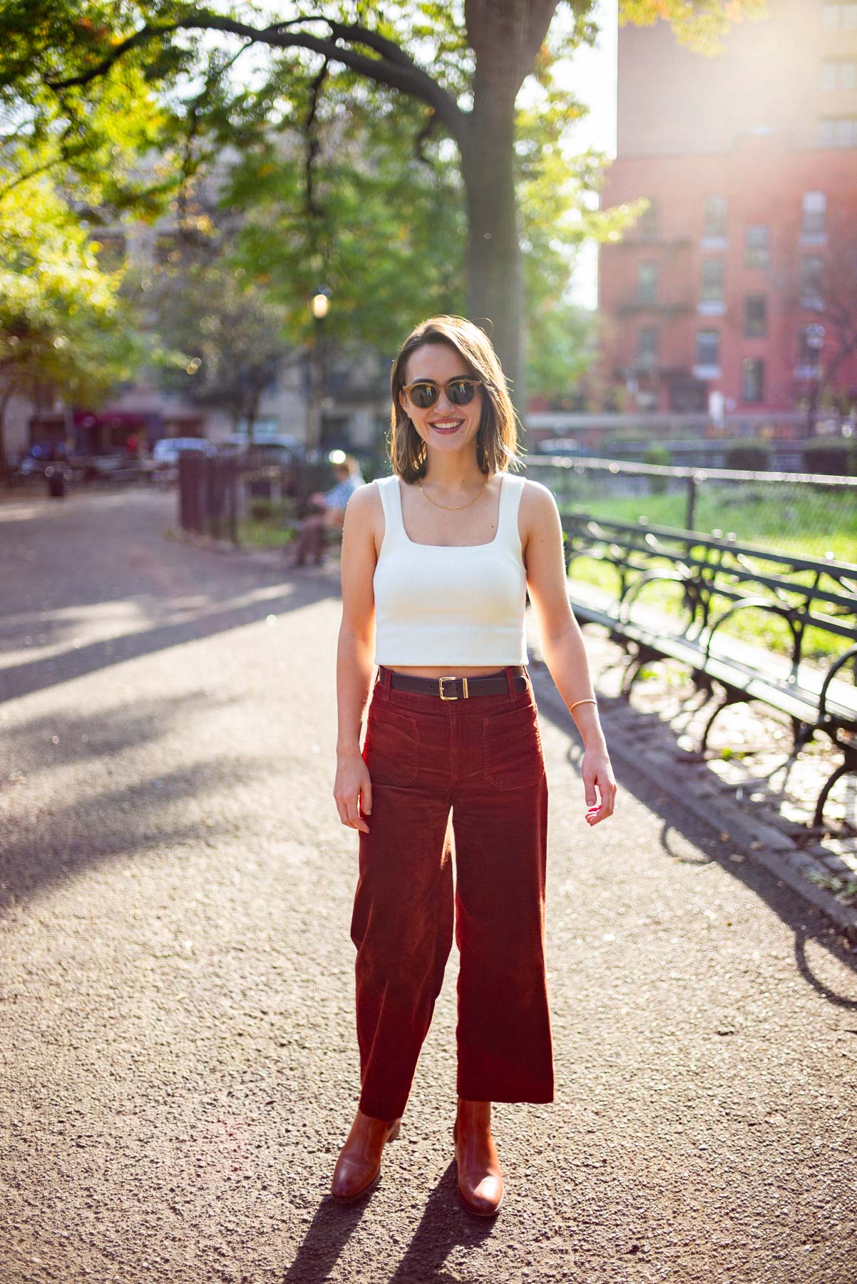 Smiling woman wearing a white tanktop and maroon wide-legged pants enjoying the sunshine at Tompkins Square Park with empty benches in the background