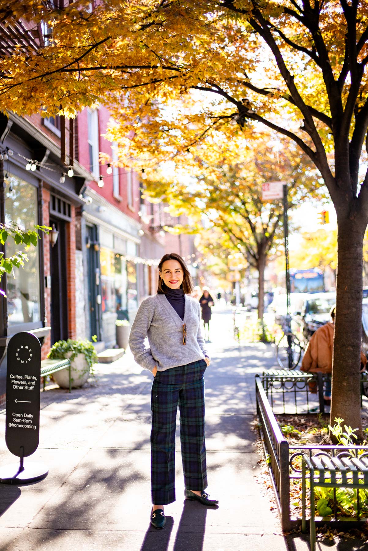Fashionable woman in a gray sweater and plaid green and blue pants standing under a sun-soaked tree in Greenpoint Brooklyn, one of the best Brooklyn neighborhoods