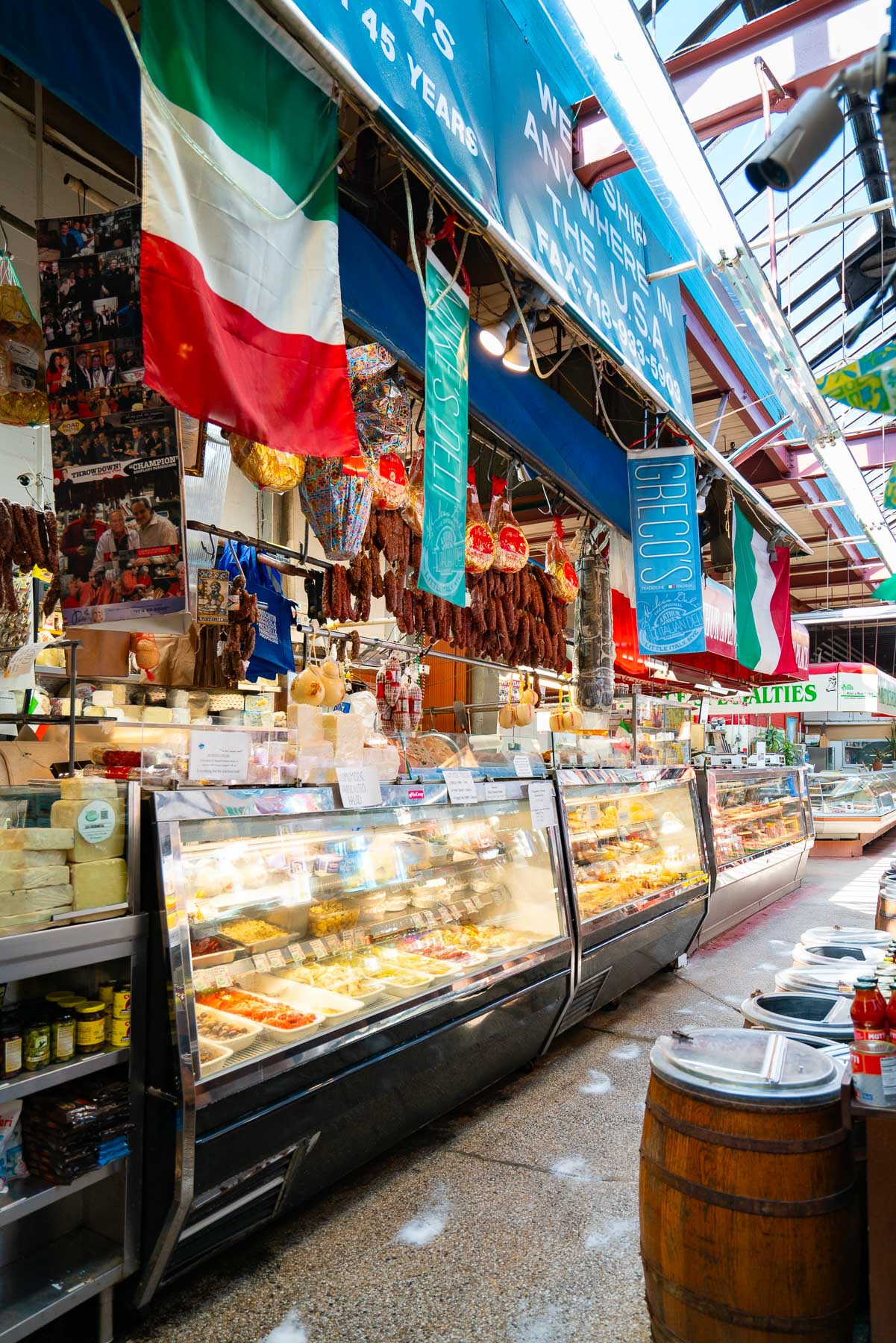 Meat and cheese counter at the Arthur Avenue Retail Market with Italian flags hanging from the ceilings and barrels of olives across the opposite end of the walkway