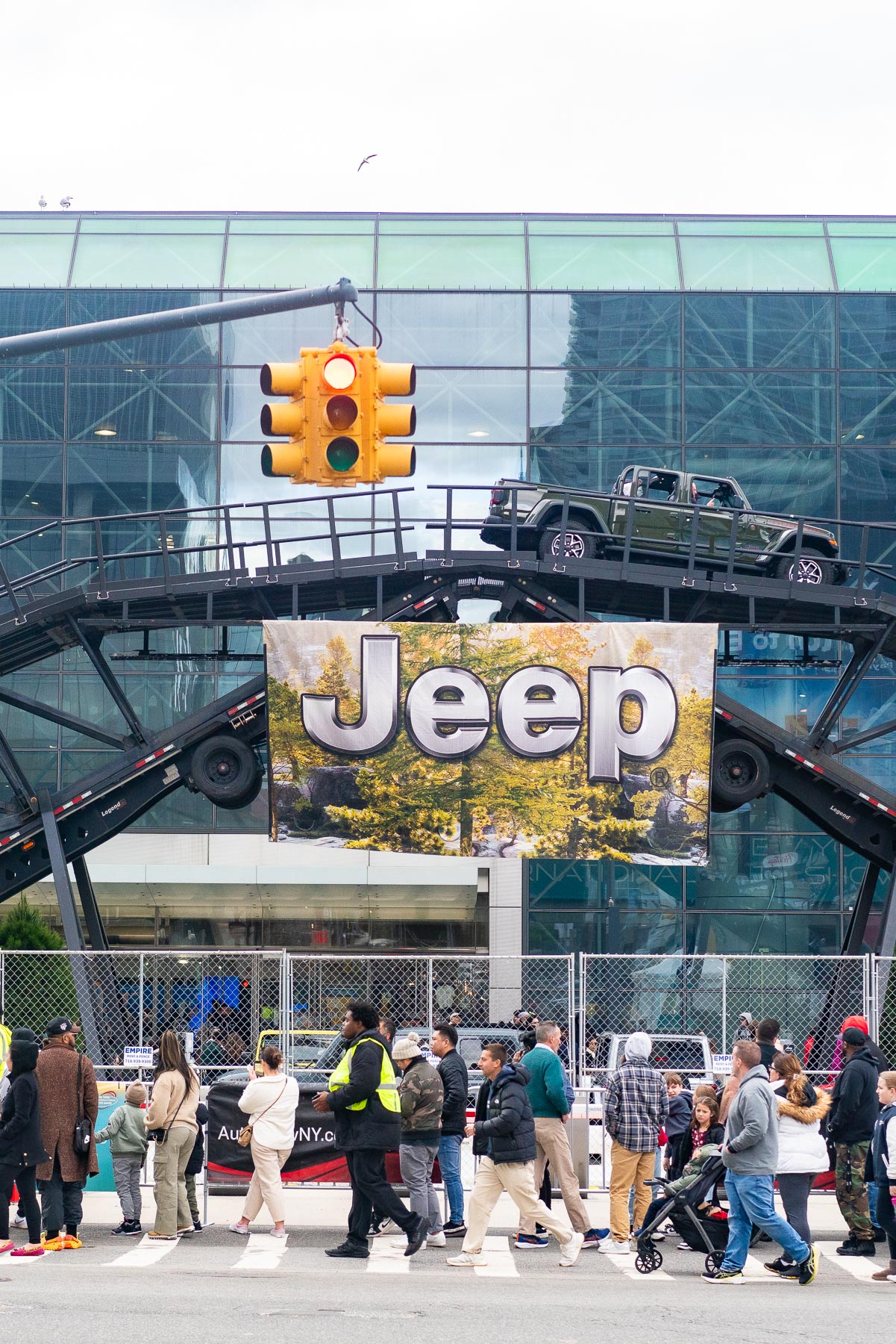 A line outside of Jacob Javits Convention Center for the New York International Auto Show as a Jeep truck drives through an elevated obstacle course in the background