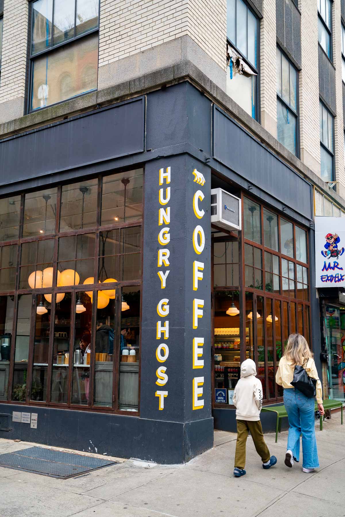 A mother and son walking past the black exterior of Hungry Ghost Coffee with yellow and white letters and big grid windows and a green bench