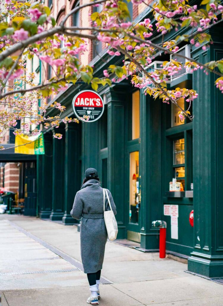 A woman in a gray pea-coat and carrying a tote bag walking past Jack's Stir Brew Coffee in Tribeca under blooming pink cherry blossoms