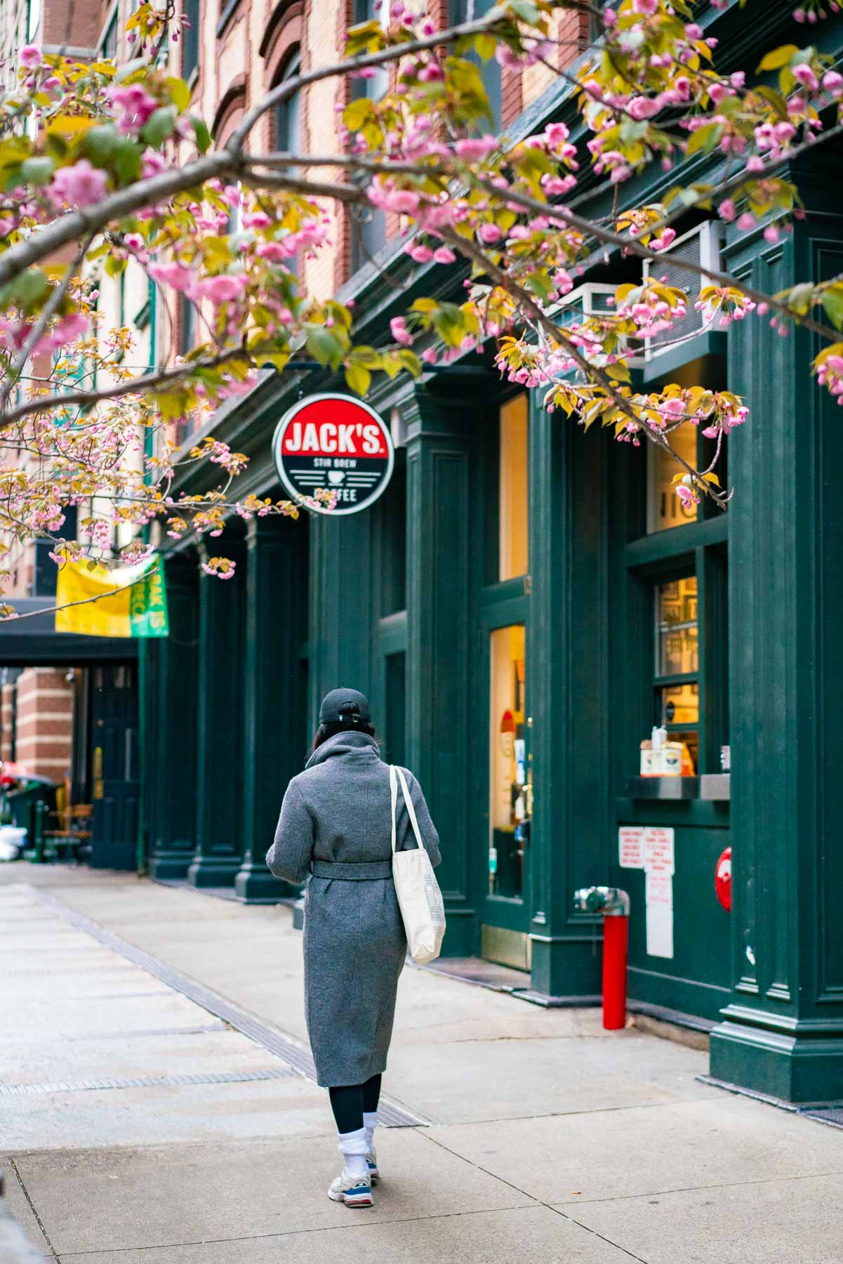 A woman in a gray pea-coat and carrying a tote bag walking past Jack's Stir Brew Coffee in Tribeca under blooming pink cherry blossoms