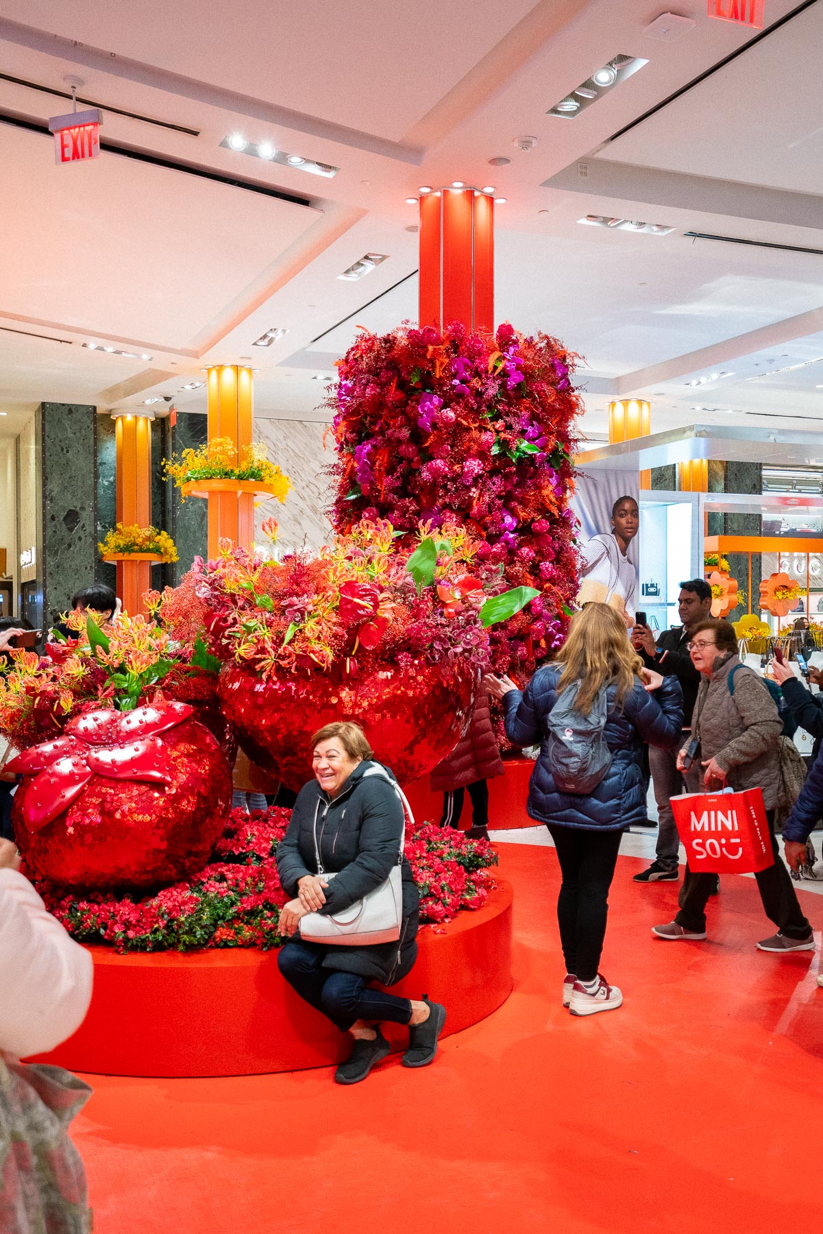 A woman sitting on a big sparkly strawberry display as other people pose and walk by at the Macy's Flower Show