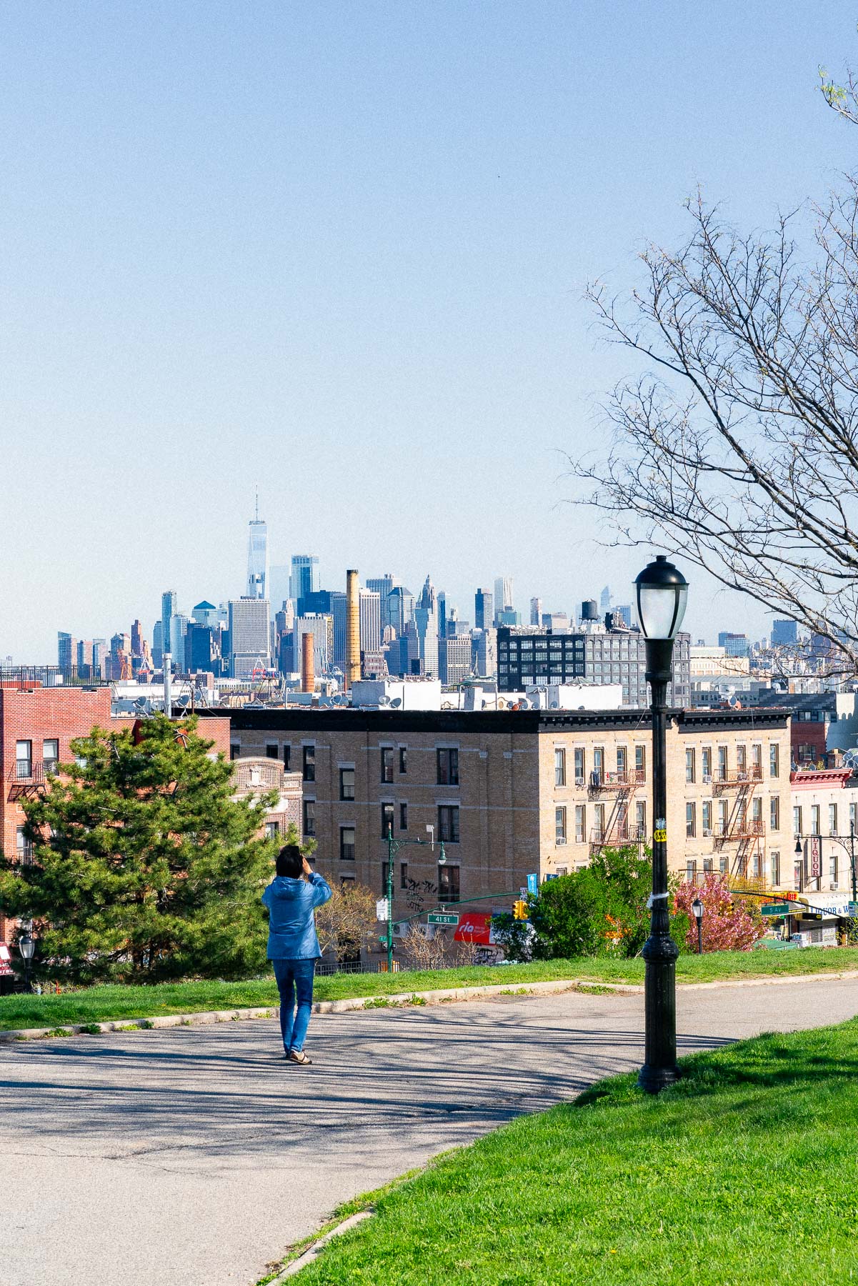 Woman in a blue jacket and jeans stopping to take a photo along the pathway of Sunset Park Brooklyn next to a lamppost, with a view of the NYC Skyline in the background