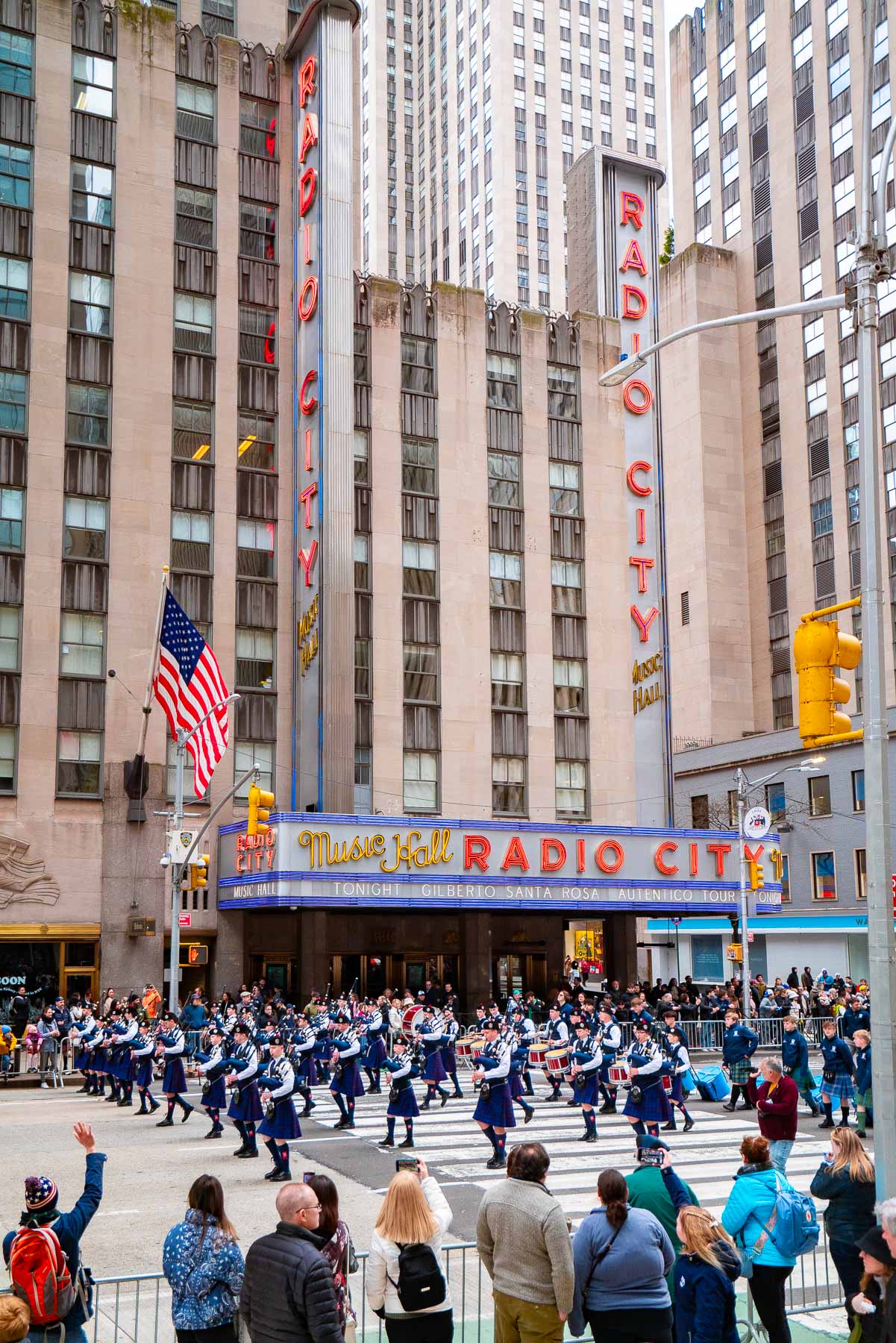 A group of bagpipe players in kilts march in front of Radio City Music Hall as a crowd of onlookers cheer them on during the Tartan Day Parade